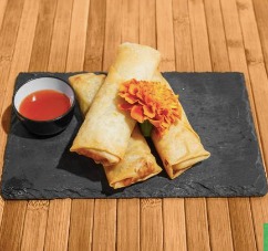 Tres rollitos de primavera crujientes sobre una piedra negra, acompañados de una pequeña porción de salsa roja y decorados con una flor. Fondo de madera clara.