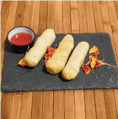 Tres rollos fritos servidos en una tabla de pizarra, acompañados de una salsa roja y decorados con pétalos de flores. Ideal para aperitivos o entradas en una comida.