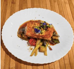 Plato gourmet de salmón glaseado servido sobre una cama de verduras asadas, incluyendo espárragos, pimientos y tomates, decorado con flores comestibles y hierbas, presentado en un plato blanco sobre una superficie de madera.